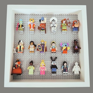 May include: A white framed display case filled with various Lego minifigures. The figures are arranged on a gray Lego baseplate. The minifigures include characters from Disney movies and other pop culture icons. The frame is white.