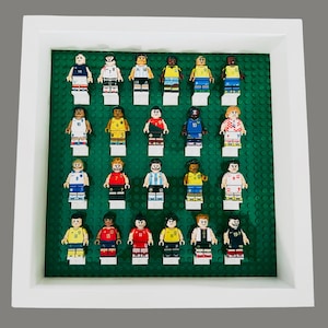 Może przedstawiać: Oprawiona ekspozycja z minifigurkami Lego, z których każda przedstawia piłkarza w różnych strojach drużynowych. Minifigurki są ułożone na zielonej podstawie w białej ramie, prezentując różnorodność kolorów i wzorów.