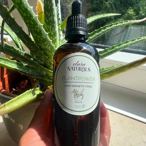 Może przedstawiać: Brązowa szklana butelka z czarnym zakraplaczem, oznaczona etykietą "clara NATURALS PLANTPOWER HAIR GROWTH TONIC". Etykieta ma zielono-biały wzór z ilustracją moździerza i tłuczka. Butelka jest trzymana przed zieloną rośliną aloesu.