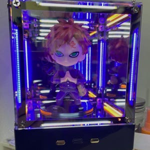 May include: A small anime-style figurine with spiky reddish-brown hair and a red kanji symbol on the forehead, wearing a dark purple outfit. The figure stands inside a rectangular, transparent display case illuminated by bright blue LED light strips.
