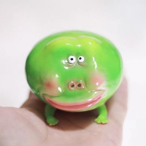 May include: A small, round, green figurine shaped like a frog with a smooth, glossy finish. It features two tiny white eyes with black pupils, a wide pink mouth, and two small nostrils. The creature has four short, stubby legs and pinkish cheeks.