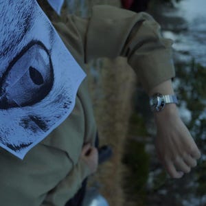 May include: A tan long-sleeved shirt featuring a large black and white graphic print of a feline eye on the left side, paired with a silver-toned wristwatch with a metallic link band worn on the wrist.