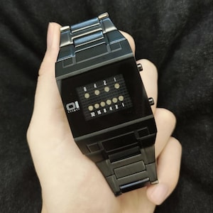 May include: A black metal binary wristwatch featuring a rectangular face with a grid display. The screen shows two rows of illuminated circular indicators with numerical labels 8, 4, 2, 1 on top and 32, 16, 8, 4, 2, 1 on the bottom. The brand logo 01 theone is visible.