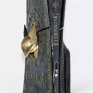 May include: A custom console cover featuring a dark, textured finish with gold-colored filigree patterns and the text ELDEN RING. A three-dimensional gold-colored winged helmet is attached to the side, with the title ELDEN RING printed on the edge.