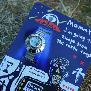 May include: A silver-toned wristwatch with a round face featuring a graphic illustration, mounted on a blue cardboard backing. The packaging displays a red UFO, a stylized face, an hourglass, and text reading "Mommy I'm going to escape from the earth tonight" and "Do you have any worries today?" with a digital clock showing 04:44.