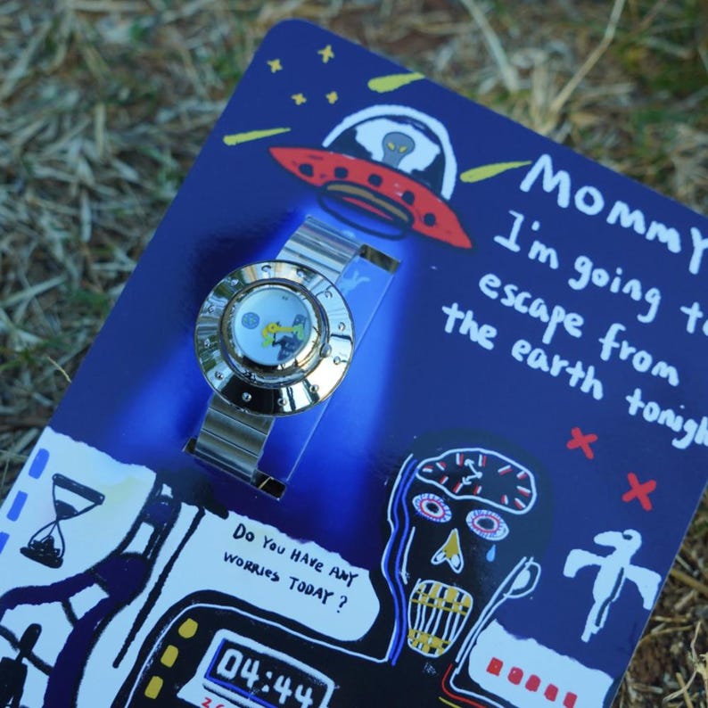 May include: A silver-toned wristwatch with a round face featuring a graphic illustration, mounted on a blue cardboard backing. The packaging displays a red UFO, a stylized face, an hourglass, and text reading "Mommy I'm going to escape from the earth tonight" and "Do you have any worries today?" with a digital clock showing 04:44.