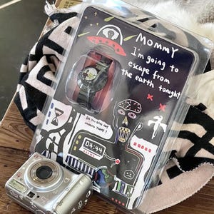 May include: A wristwatch in clear plastic packaging featuring a black band and a round face. The packaging displays a red UFO, a digital clock reading 04:44, and the text: Mommy I'm going to escape from the earth tonight and Do you have any worries today?