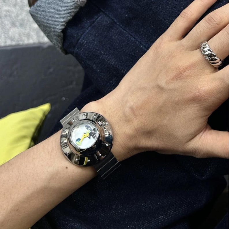 May include: A silver-toned wristwatch with a wide, circular, polished metal bezel featuring small decorative studs around the edge. The watch face has a white background with a bright yellow hand. A thick, twisted silver-toned ring is worn on the finger.
