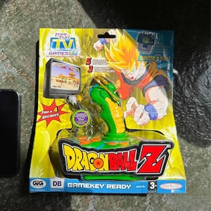 Op de afbeelding: Dragon Ball Z Gamekey Ready plug-and-play tv-spel. De verpakking heeft een groen en geel kleurenschema met het Dragon Ball Z-logo. De gamecontroller is groen en oranje, met een karakterontwerp. Het pakket bevat de tekst "Fino a 4 giocatori" en "Gamekey Ready".