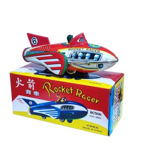 May include: A vintage tin toy rocket racer in red, white, and silver, with the number 6 on the fin. The toy sits atop a red and yellow box with the words "Rocket Racer" and a cartoon rocket ship illustration.