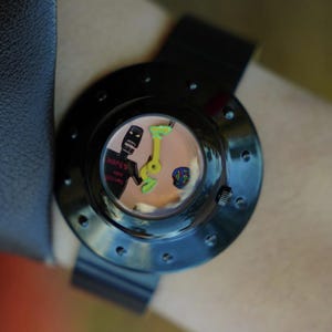 May include: A black wristwatch with a circular case featuring a series of small indentations around the bezel. The watch face displays a light tan background with a black figure, a small colorful globe, and the text Hopes and Dreams in red.