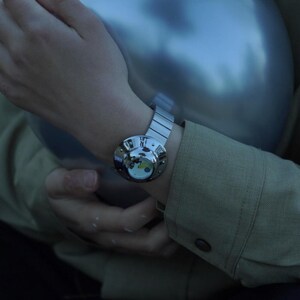 May include: A silver-toned wristwatch featuring a circular, polished metallic case with a domed glass cover. The watch face displays a white background with small yellow and blue graphic accents. It includes a flexible, segmented metal link band.