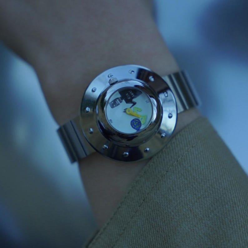 May include: A silver-toned wristwatch featuring a wide, polished circular bezel with small decorative rivets. The watch face is white with a graphic illustration, yellow hands, and a metallic band. The text RUN AWAY is printed on the watch face.