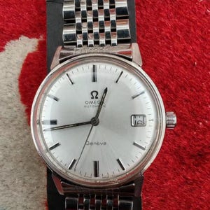 Pode incluir: Um relógio de pulso automático Omega Genève prateado com mostrador redondo branco, marcadores de hora pretos, janela de data na posição das três horas e uma pulseira de elos de aço inoxidável com design de várias fileiras.