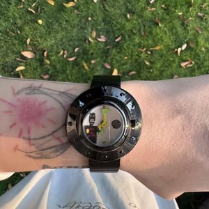 May include: A black wristwatch with a circular case and a metallic band. The watch face features a white background with a stylized black figure, a small green graphic, and bright yellow hands. The bezel is glossy black with small circular indentations.
