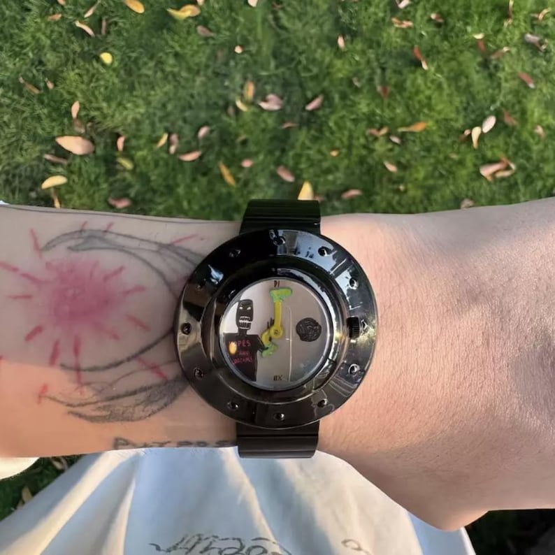 May include: A black wristwatch with a circular case and a metallic band. The watch face features a white background with a stylized black figure, a small green graphic, and bright yellow hands. The bezel is glossy black with small circular indentations.