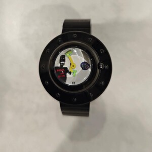 May include: A black wristwatch with a circular frame featuring a white dial. The dial displays a black figure with the red text "HOPES AND DREAMS," a yellow hand, and a small multicolored circular graphic. The watch has a black band and a bezel with dots.