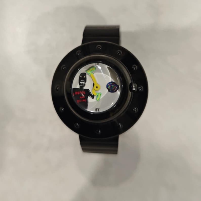 May include: A black wristwatch with a circular frame featuring a white dial. The dial displays a black figure with the red text "HOPES AND DREAMS," a yellow hand, and a small multicolored circular graphic. The watch has a black band and a bezel with dots.