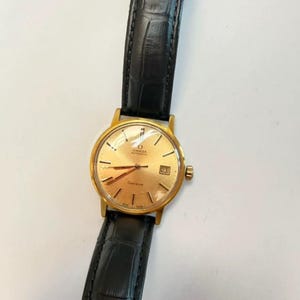Pode incluir: Um relógio de pulso automático Omega Geneve com caixa redonda em tom dourado, mostrador cor de champanhe, marcadores de hora dourados e uma janela de data na posição das três horas. O relógio possui uma pulseira de couro preta texturizada.