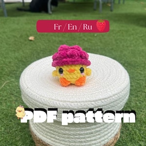Peut inclure: Une petite figurine de poussin crocheté jaune portant un chapeau texturé en forme de framboise rose foncé. Le poussin a des yeux en perles noires, un petit bec orange et des pattes orange. Le texte "Fr / En / Ru" et "PDF pattern" est visible.