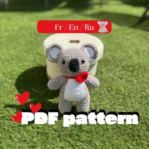 Peut inclure: Un koala en crochet avec une fourrure gris clair, un ventre blanc, des yeux noirs et un nez noir. Le koala porte un petit cœur rouge au niveau du cou. Une bannière rouge en haut affiche le texte Fr / En / Ru, et le texte PDF pattern est visible en bas.