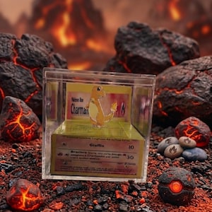 Può includere: Una teca in acrilico trasparente contenente una carta collezionabile di Charmander su sfondo giallo. La carta presenta l'illustrazione del personaggio, il nome Charmander e testo in italiano tra cui Graffio 10 e Braciere 30 come oggetto da collezione.