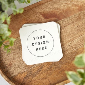 May include: Two white square coasters with rounded corners on a wooden tray. The top coaster has the text "Your Design Here" in a circle.