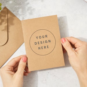 Maquette de carte de voeux kraft marron avec enveloppe et modèle à la main. Soleil tacheté. Carte d'anniversaire tenue à la main. Invitation de mariage. Mise à plat.. Jpeg uniquement