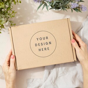 Puede incluir: Una caja de cartón marrón con el texto "YOUR DESIGN HERE" en un círculo en la parte delantera. La caja está siendo sostenida por las manos de una persona.