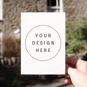 New home greeting card mockup with models hand. Landscape outdoors mock up with cosy cottage. Jpeg only.