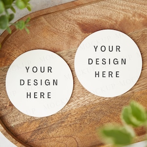 Peut inclure: Deux dessous-de-verre ronds blancs avec le texte "YOUR DESIGN HERE" sur un plateau en bois. Le plateau est entouré de plantes vertes.