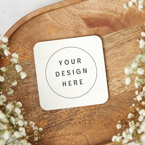 May include: A white square coaster with a circle in the center that says "Your Design Here" on a wooden tray with white flowers.