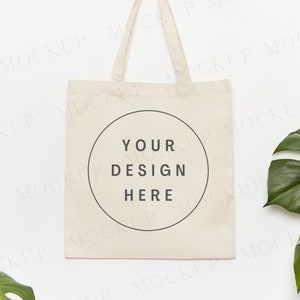 May include: A white canvas tote bag with a circle in the center that says "Your Design Here". The bag is hanging on a white wall with green monstera leaves on either side.