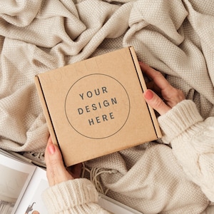 Puede incluir: Una caja de cartón marrón con el texto "YOUR DESIGN HERE" en un círculo en la parte delantera. La caja está siendo sostenida por una persona que lleva un suéter blanco.