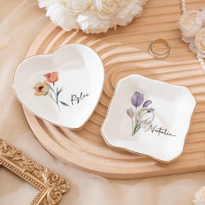 Könnte beinhalten: Zwei weiße Keramik-Ringschalen mit goldenem Rand. Eine ist herzförmig, die andere quadratisch. Beide haben Blumendesigns und personalisierte Namen. Ein Strauß weißer Blumen und eine Perlenkette befinden sich im Hintergrund.