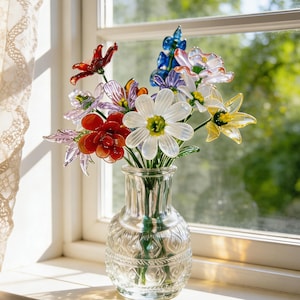 May include: A clear glass vase filled with colorful glass flowers sits on a windowsill. The flowers are various colors, including red, white, yellow, and blue. The vase is in front of a window with a lace curtain.