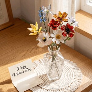 Pode incluir: Um vaso de vidro transparente com padrão geométrico contém um buquê de flores de vidro coloridas em tons de amarelo, azul, vermelho, laranja e branco. Ao lado do vaso, há um cartão branco dobrado com o texto impresso Happy Mother's Day.