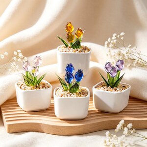 May include: Four small white square ceramic pots each containing a glass tulip flower arrangement. The tulips are in shades of yellow, blue, pink, and purple. The pots are filled with small pebbles and green glass leaves.