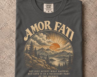 Könnte beinhalten: Ein anthrazitfarbenes T-Shirt mit Rundhalsausschnitt und einem Grafikdruck einer Berglandschaft bei Sonnenuntergang. Der Text AMOR FATI ist über dem Motiv gewölbt, darunter steht das Zitat NOT JUST ACCEPT WHAT HAPPENS BUT LOVE IT AS A NECESSARY PART OF THE JOURNEY.