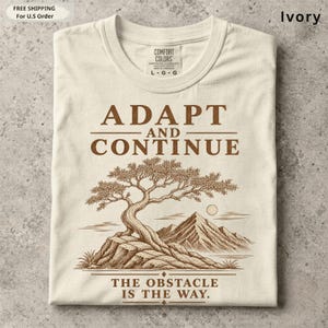 K&ouml;nnte beinhalten: Ein elfenbeinfarbenes T-Shirt mit einem braunen Grafikdruck eines Bonsai-Baums auf einem Felsvorsprung mit Bergen im Hintergrund. Der Text ADAPT AND CONTINUE steht &uuml;ber dem Baum und THE OBSTACLE IS THE WAY darunter.