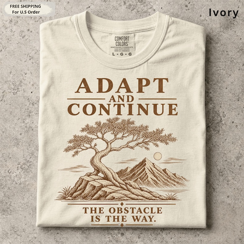 K&ouml;nnte beinhalten: Ein elfenbeinfarbenes T-Shirt mit einem braunen Grafikdruck eines Bonsai-Baums auf einem Felsvorsprung mit Bergen im Hintergrund. Der Text ADAPT AND CONTINUE steht &uuml;ber dem Baum und THE OBSTACLE IS THE WAY darunter.