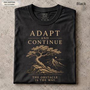 K&ouml;nnte beinhalten: Ein schwarzes T-Shirt mit Rundhalsausschnitt und goldenem Grafikdruck. Das Design zeigt den Text ADAPT AND CONTINUE, einen stilisierten Bonsai auf einem Felsen, ein Gebirge und den Text THE OBSTACLE IS THE WAY.