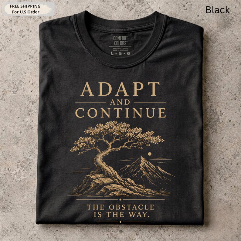 K&ouml;nnte beinhalten: Ein schwarzes T-Shirt mit Rundhalsausschnitt und goldenem Grafikdruck. Das Design zeigt den Text ADAPT AND CONTINUE, einen stilisierten Bonsai auf einem Felsen, ein Gebirge und den Text THE OBSTACLE IS THE WAY.