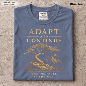 K&ouml;nnte beinhalten: Ein blaues T-Shirt mit einer goldenen Grafik eines Baumes auf einer felsigen Klippe, Bergen und einer Sonne im Hintergrund. Der Text ADAPT AND CONTINUE steht &uuml;ber dem Baum und THE OBSTACLE IS THE WAY unter dem Baum.