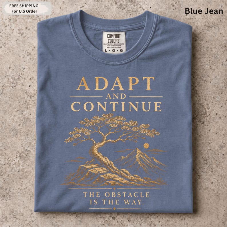 K&ouml;nnte beinhalten: Ein blaues T-Shirt mit einer goldenen Grafik eines Baumes auf einer felsigen Klippe, Bergen und einer Sonne im Hintergrund. Der Text ADAPT AND CONTINUE steht &uuml;ber dem Baum und THE OBSTACLE IS THE WAY unter dem Baum.