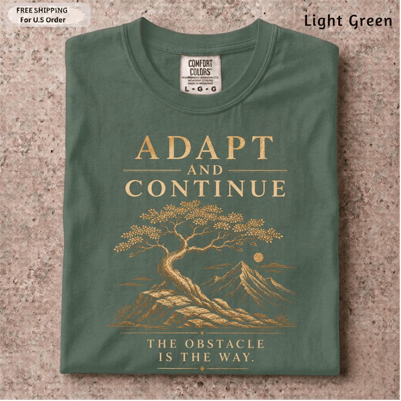 K&ouml;nnte beinhalten: Ein hellgr&uuml;nes T-Shirt mit einer goldenen Grafik eines Bonsai-Baums auf einem Felsvorsprung und Bergen im Hintergrund. Der Text ADAPT AND CONTINUE steht &uuml;ber dem Baum und THE OBSTACLE IS THE WAY unter dem Baumdesign.