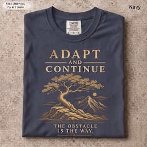 K&ouml;nnte beinhalten: Ein marineblaues T-Shirt mit einer goldenen Grafik eines Baumes auf einem Felsvorsprung vor Bergen. Der Text lautet ADAPT AND CONTINUE und THE OBSTACLE IS THE WAY. Das T-Shirt hat ein Nackenetikett der Marke Comfort Colors.