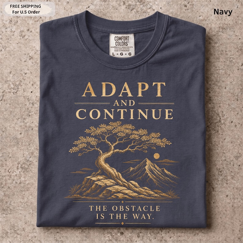 K&ouml;nnte beinhalten: Ein marineblaues T-Shirt mit einer goldenen Grafik eines Baumes auf einem Felsvorsprung vor Bergen. Der Text lautet ADAPT AND CONTINUE und THE OBSTACLE IS THE WAY. Das T-Shirt hat ein Nackenetikett der Marke Comfort Colors.