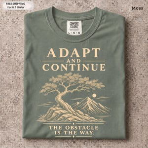 K&ouml;nnte beinhalten: Ein moosgr&uuml;nes T-Shirt mit einer cremefarbenen Grafik eines Bonsai-Baums auf einem Felsen und Bergen im Hintergrund. Der Text ADAPT AND CONTINUE steht &uuml;ber dem Baum und THE OBSTACLE IS THE WAY steht unter dem Baum.