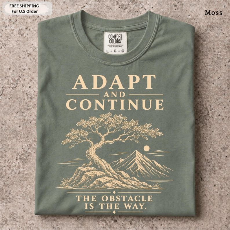 K&ouml;nnte beinhalten: Ein moosgr&uuml;nes T-Shirt mit einer cremefarbenen Grafik eines Bonsai-Baums auf einem Felsen und Bergen im Hintergrund. Der Text ADAPT AND CONTINUE steht &uuml;ber dem Baum und THE OBSTACLE IS THE WAY steht unter dem Baum.